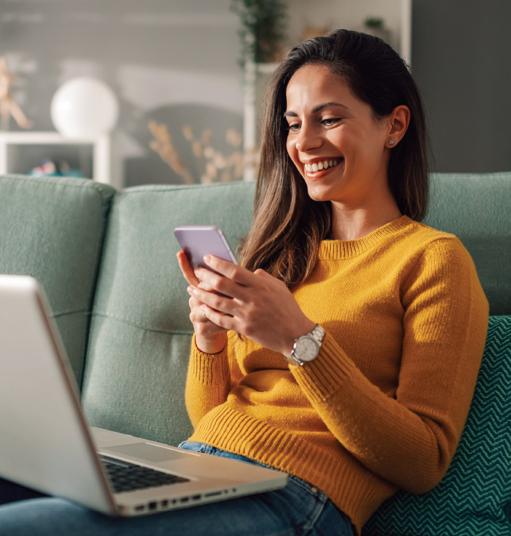 A young woman looking at her phone while sitting on her couch, smiling and happy